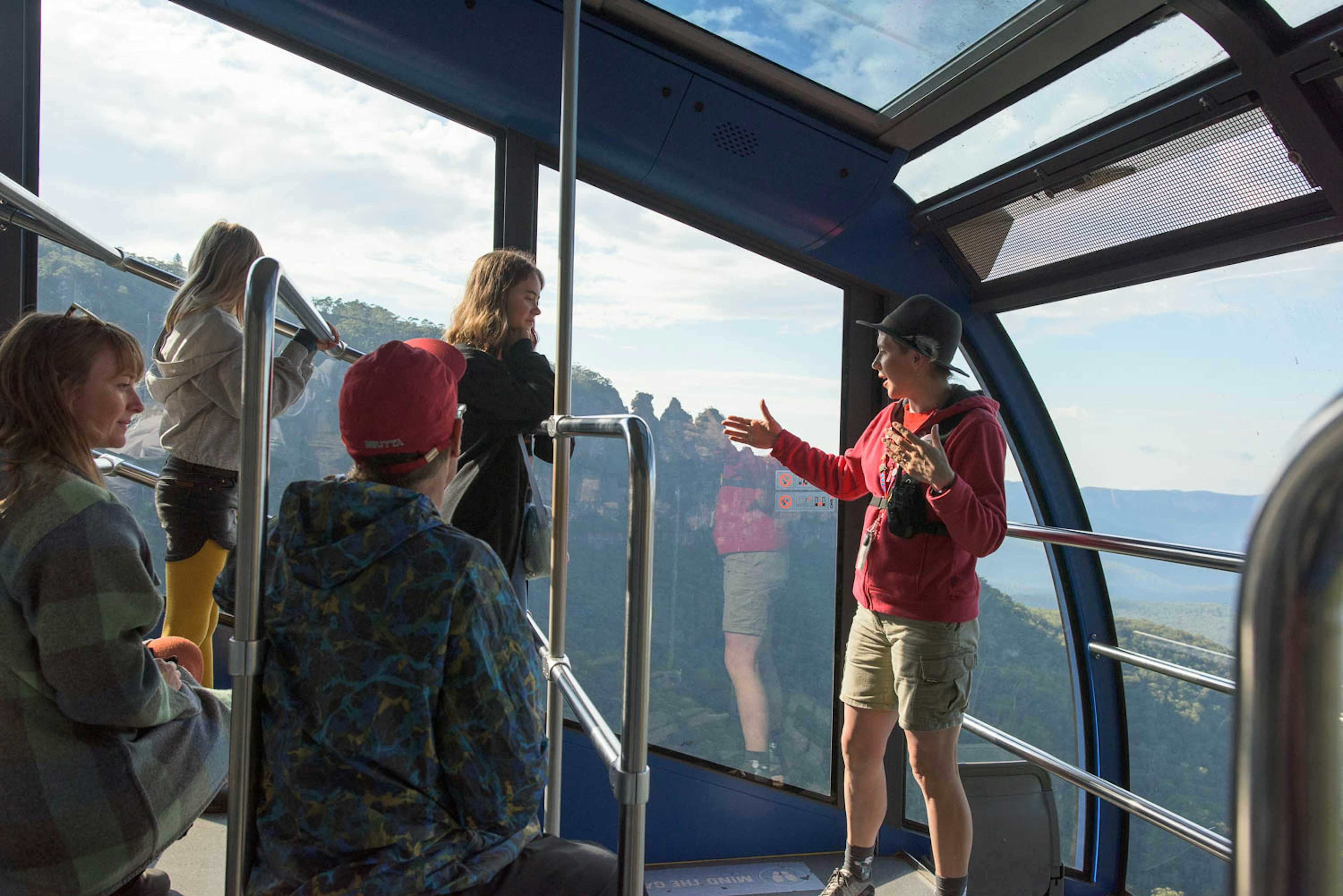 A guide is talking to a group of people inside the Cableway cable car with a view of the Three Sisters.  