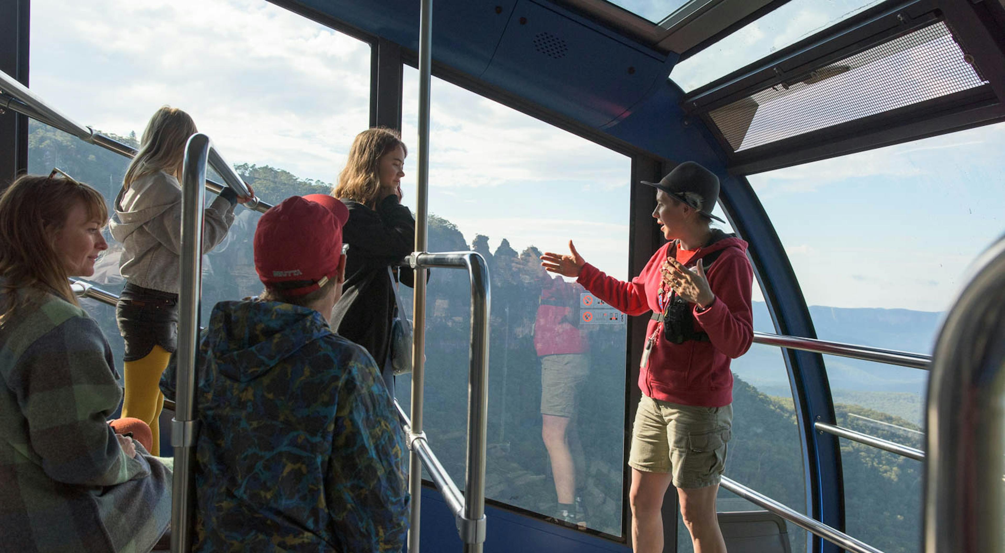 A guide is talking to a group of people inside the Cableway cable car with a view of the Three Sisters.  