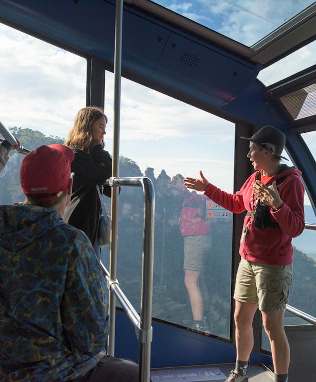 A group of people enjoy a scenic view from inside a glass-walled cable car. One person gestures animatedly while others look on. The background shows a mountain landscape under a partly cloudy sky.