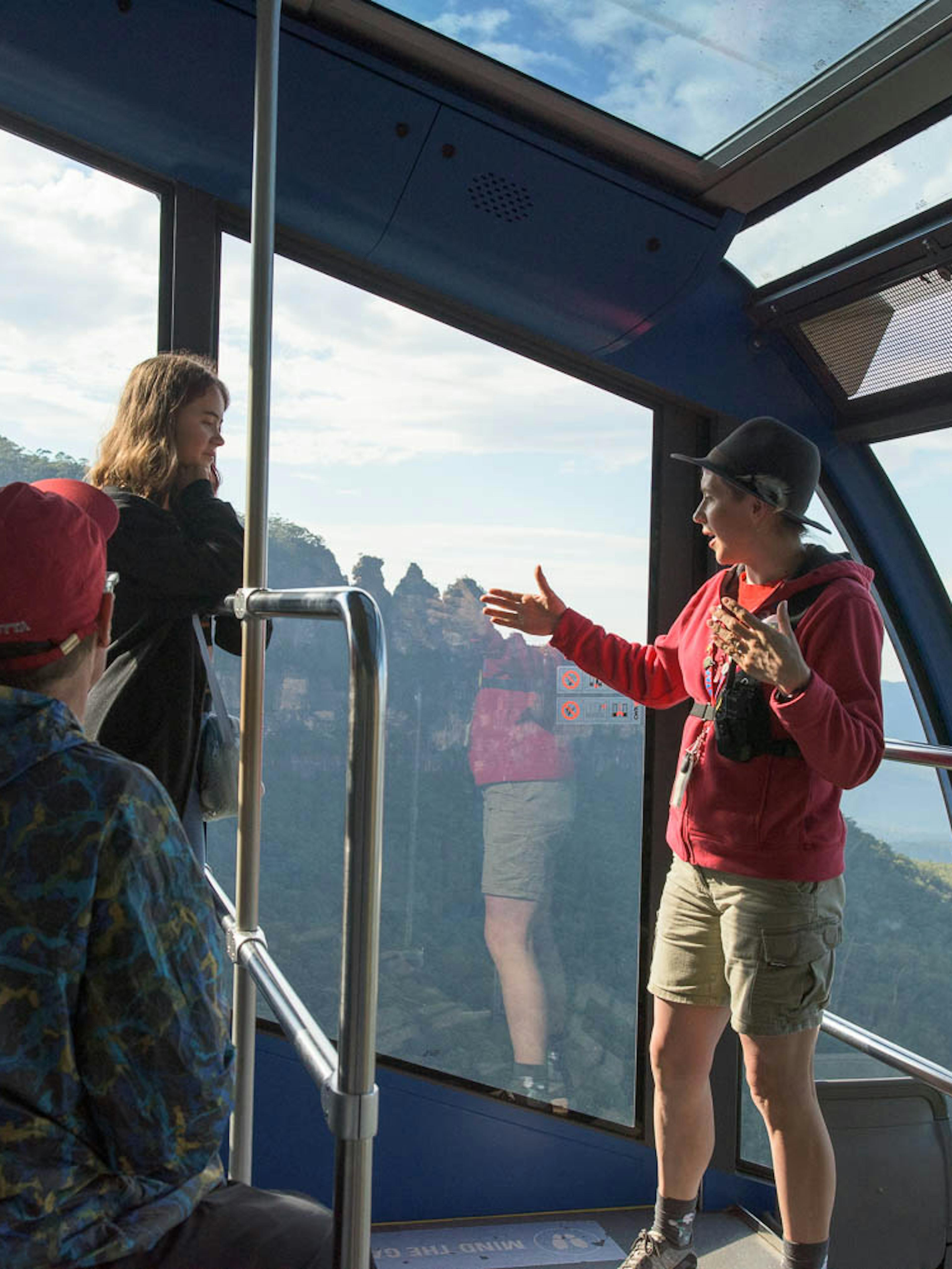 A guide is talking to a group of people inside the Cableway cable car with a view of the Three Sisters.  