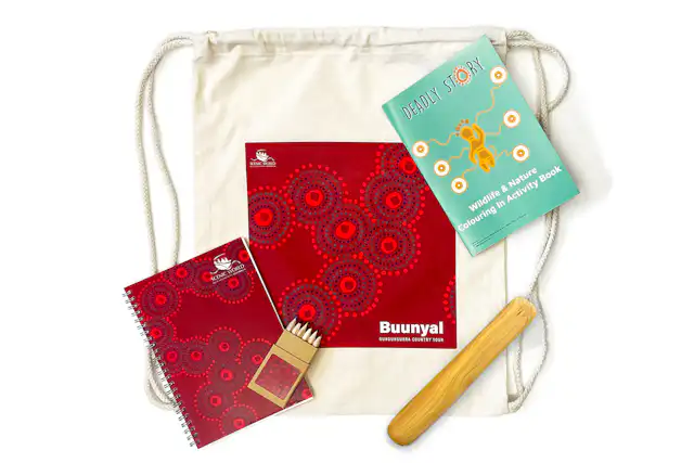 A drawstring bag containing a red spiral-bound notebook with circular patterns, a pack of coloured pencils, a wildlife and environment activity book, and a wooden stick.