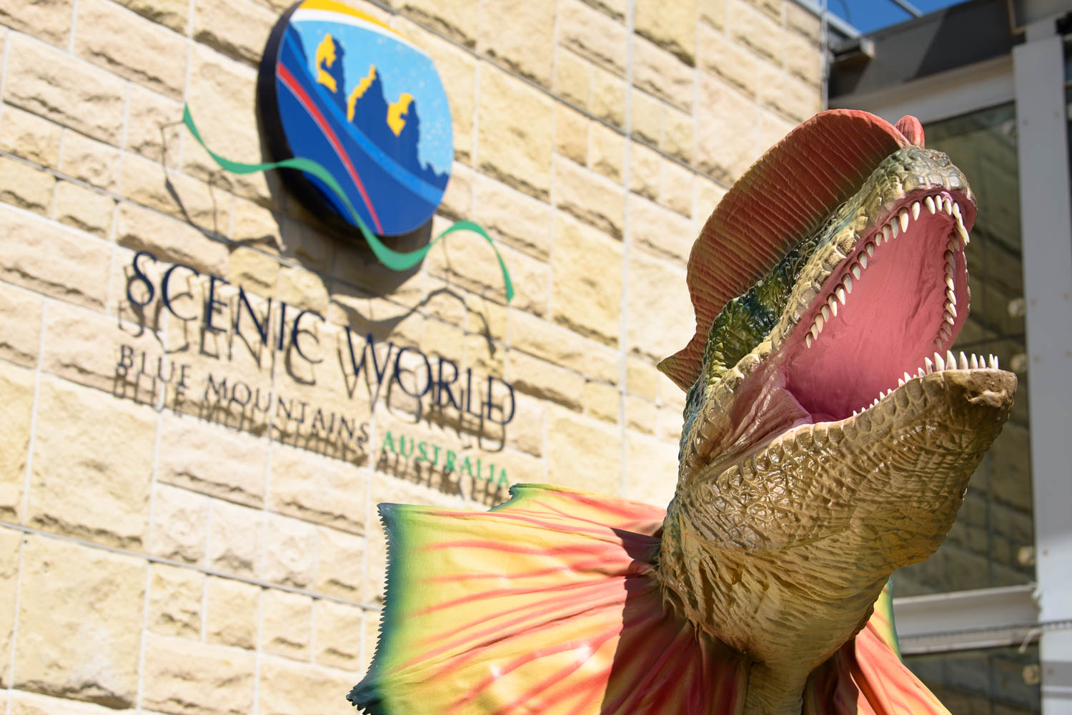 A large dinosaur with an open mouth and vibrant frill stands in front of a stone wall. An oval sign reading Scenic World Blue Mountains Australia is mounted on the wall behind the statue.