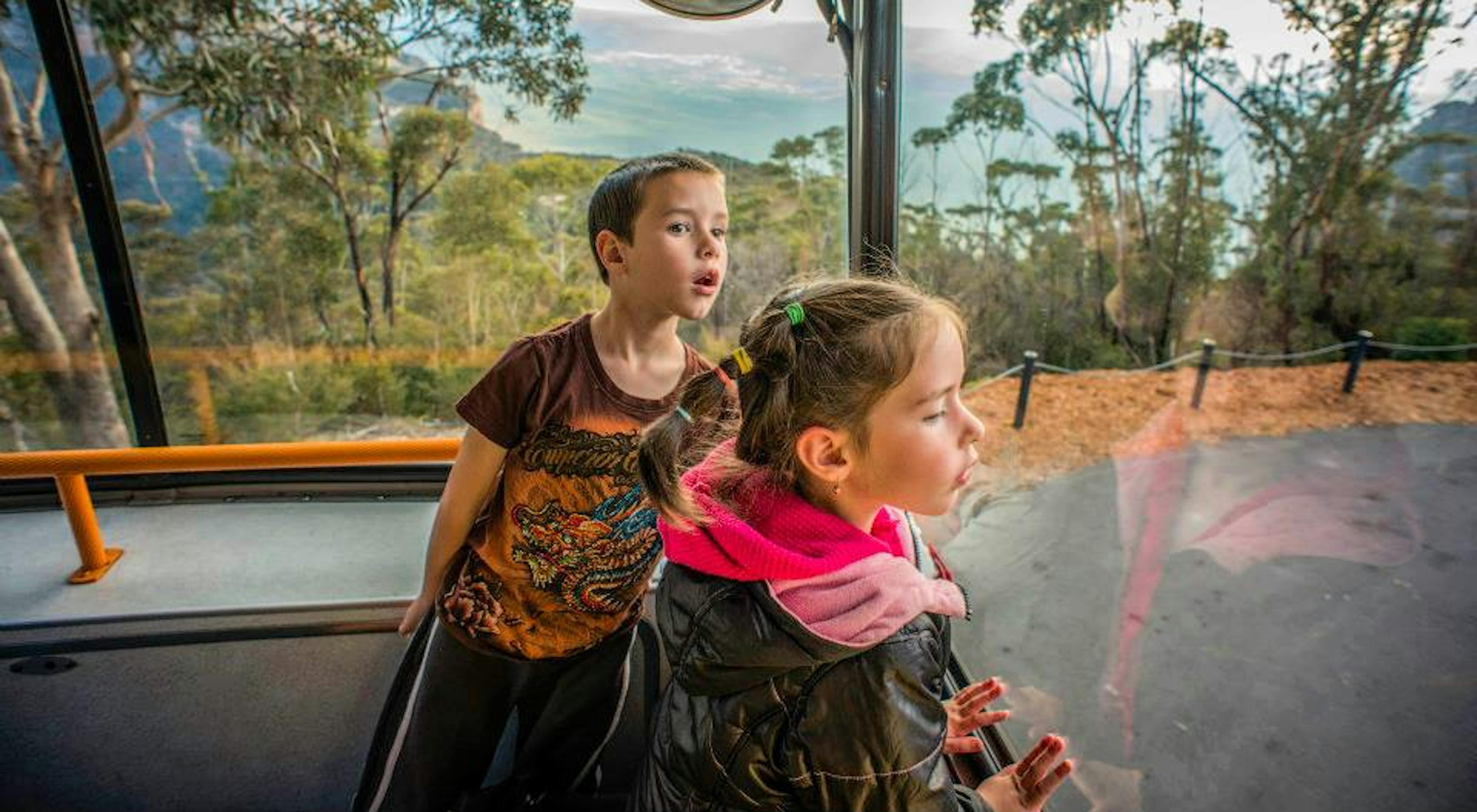 Two children eagerly look out the front window of a bus as it travels through a scenic area with trees and a distant view of mountains. A convex mirror above them reflects a portion of the bus interior.