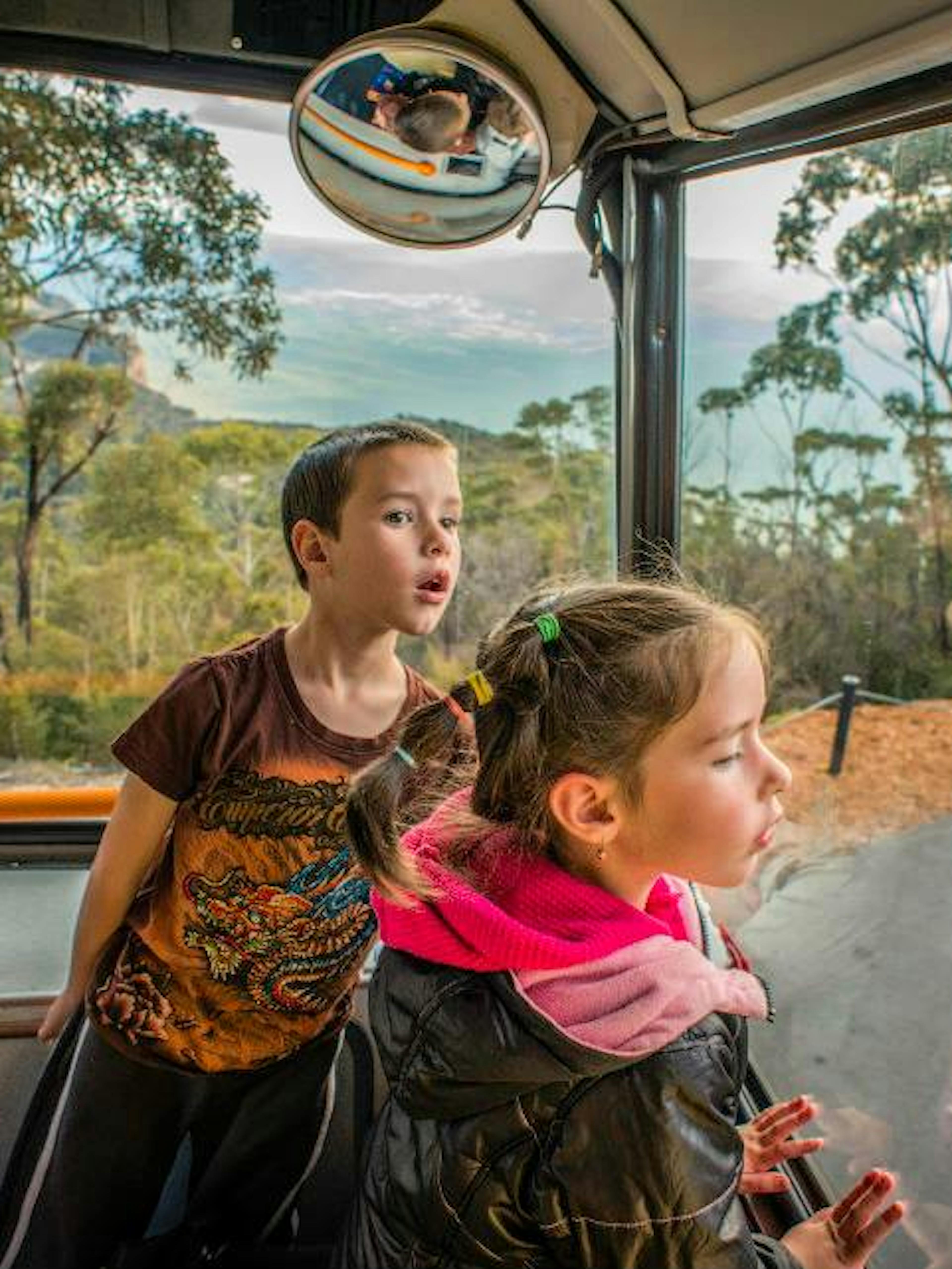 Two children eagerly look out the front window of a bus as it travels through a scenic area with trees and a distant view of mountains. A convex mirror above them reflects a portion of the bus interior.