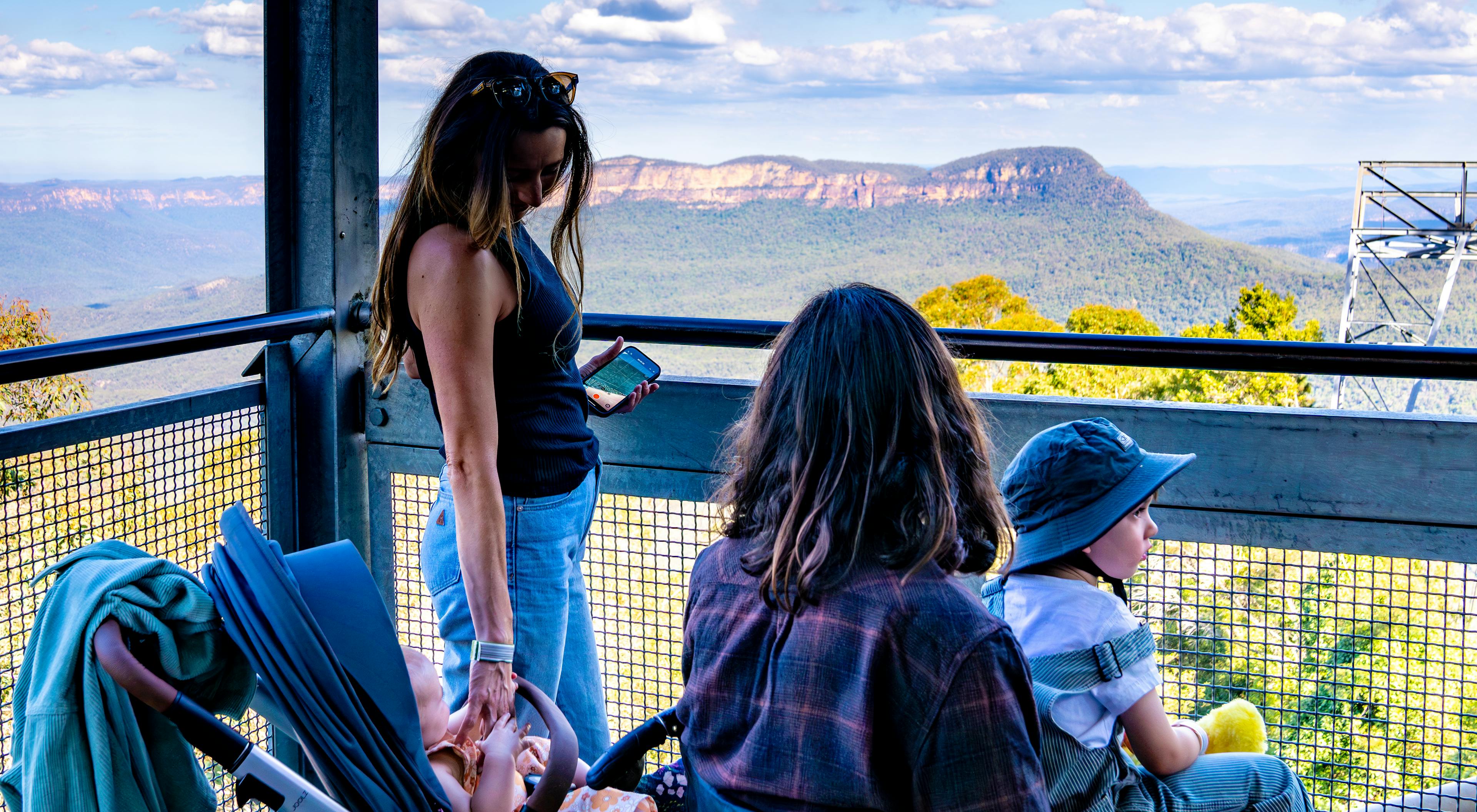 A woman stands on a lookout deck overlooking a valley and with a baby in a pram beside her. A man and a child wearing a sunhat and holding a toy sit nearby, enjoying the scenic view under a cloudy blue sky. the man is in a wheelchair 