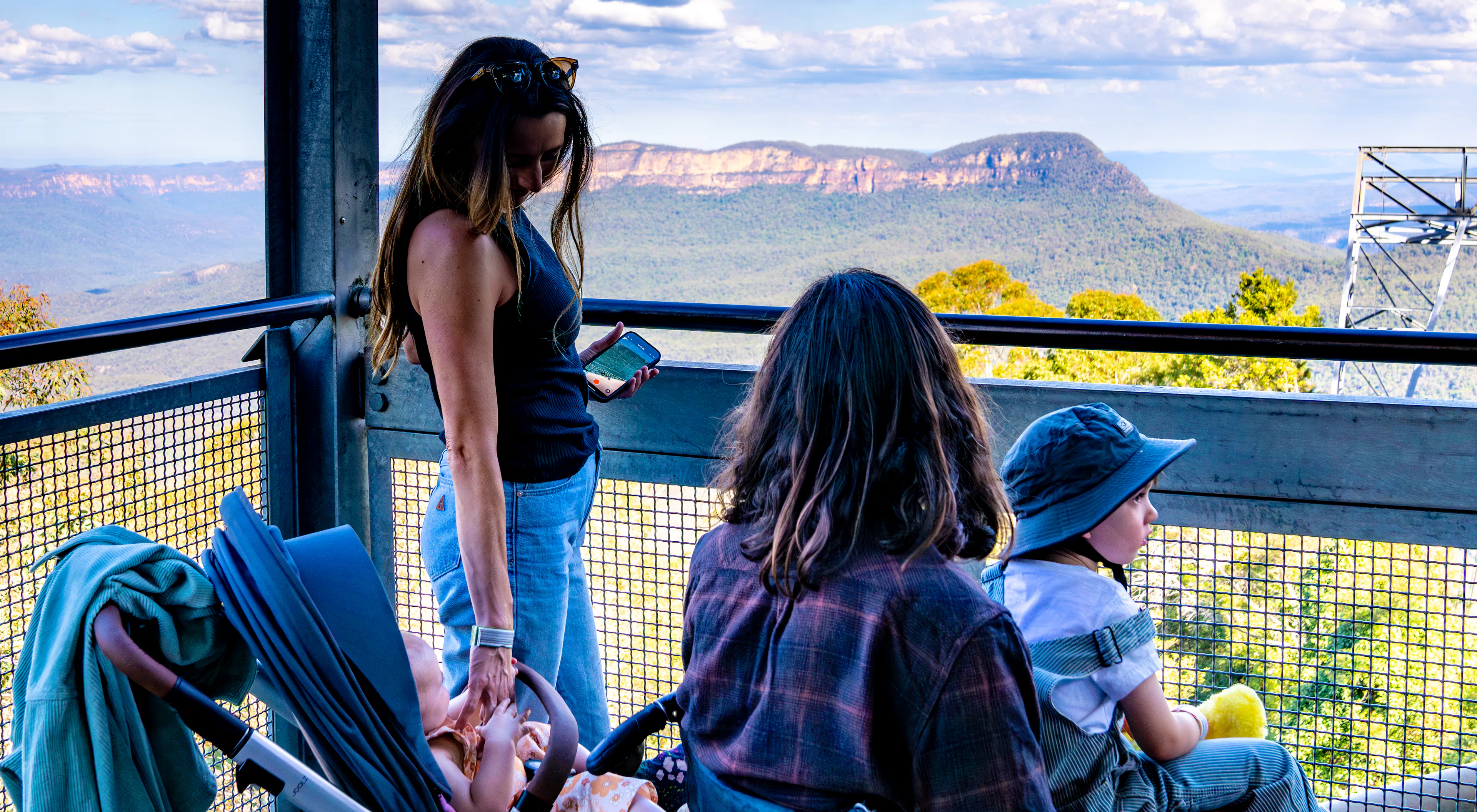 A woman stands on a lookout deck overlooking a mountainous landscape with a stroller beside her. A man with long hair and a child wearing a sunhat and holding a toy sit nearby, enjoying the scenic view under a cloudy blue sky.
