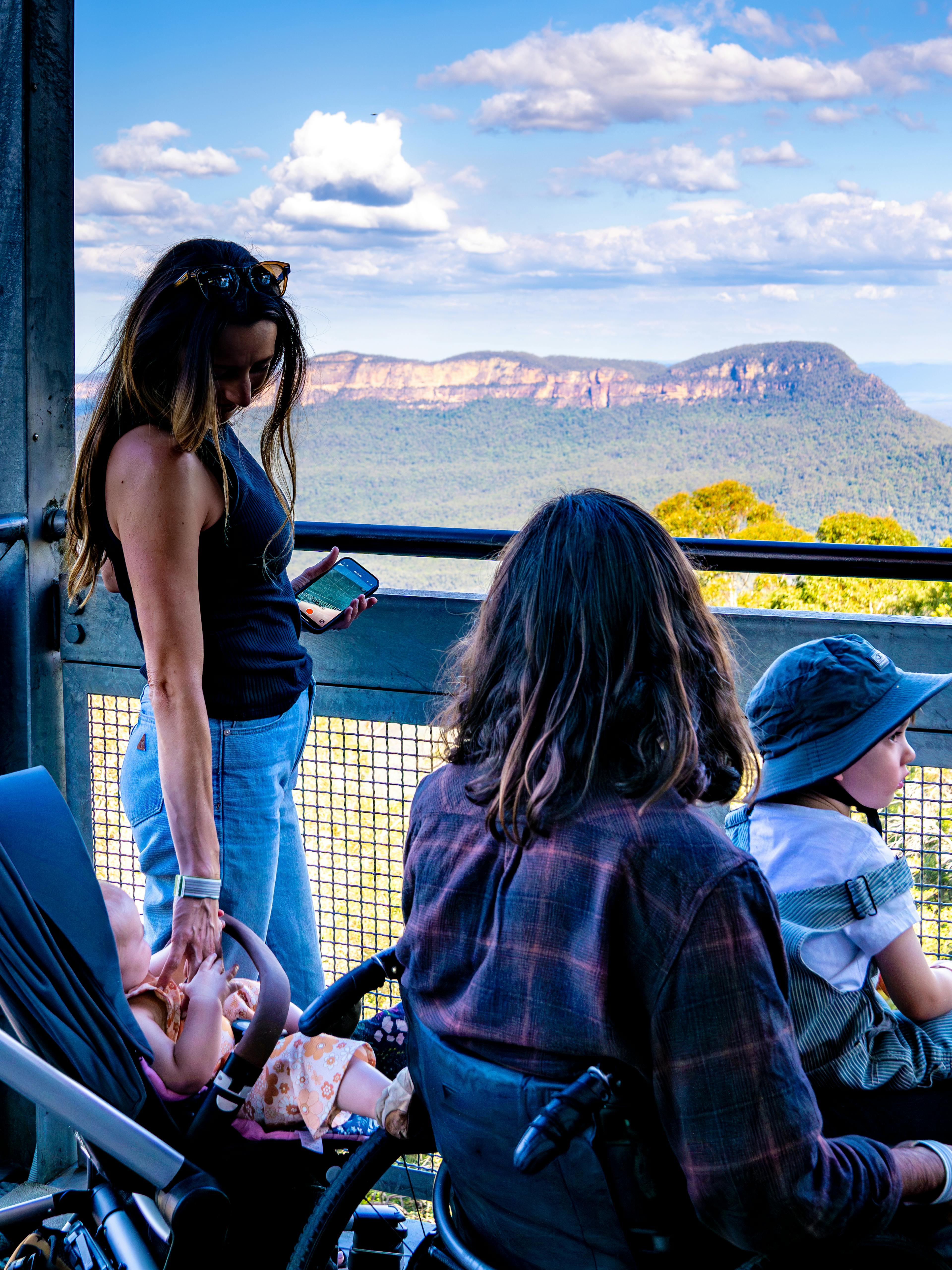 A woman stands on a lookout deck overlooking a mountainous landscape with a stroller beside her. A man with long hair and a child wearing a sunhat and holding a toy sit nearby, enjoying the scenic view under a cloudy blue sky.