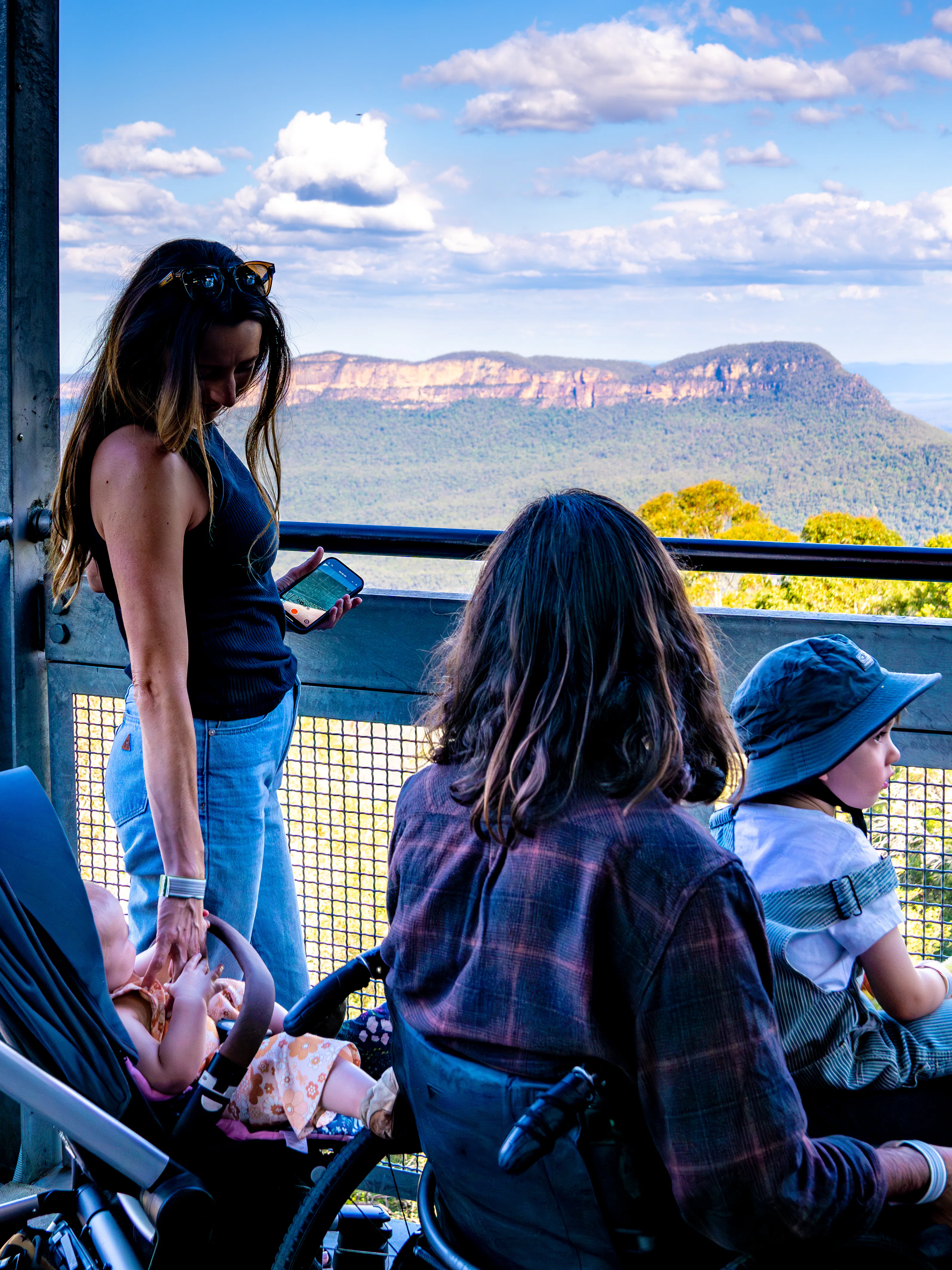 A woman stands on a lookout deck overlooking a mountainous landscape with a stroller beside her. A man with long hair and a child wearing a sunhat and holding a toy sit nearby, enjoying the scenic view under a cloudy blue sky.