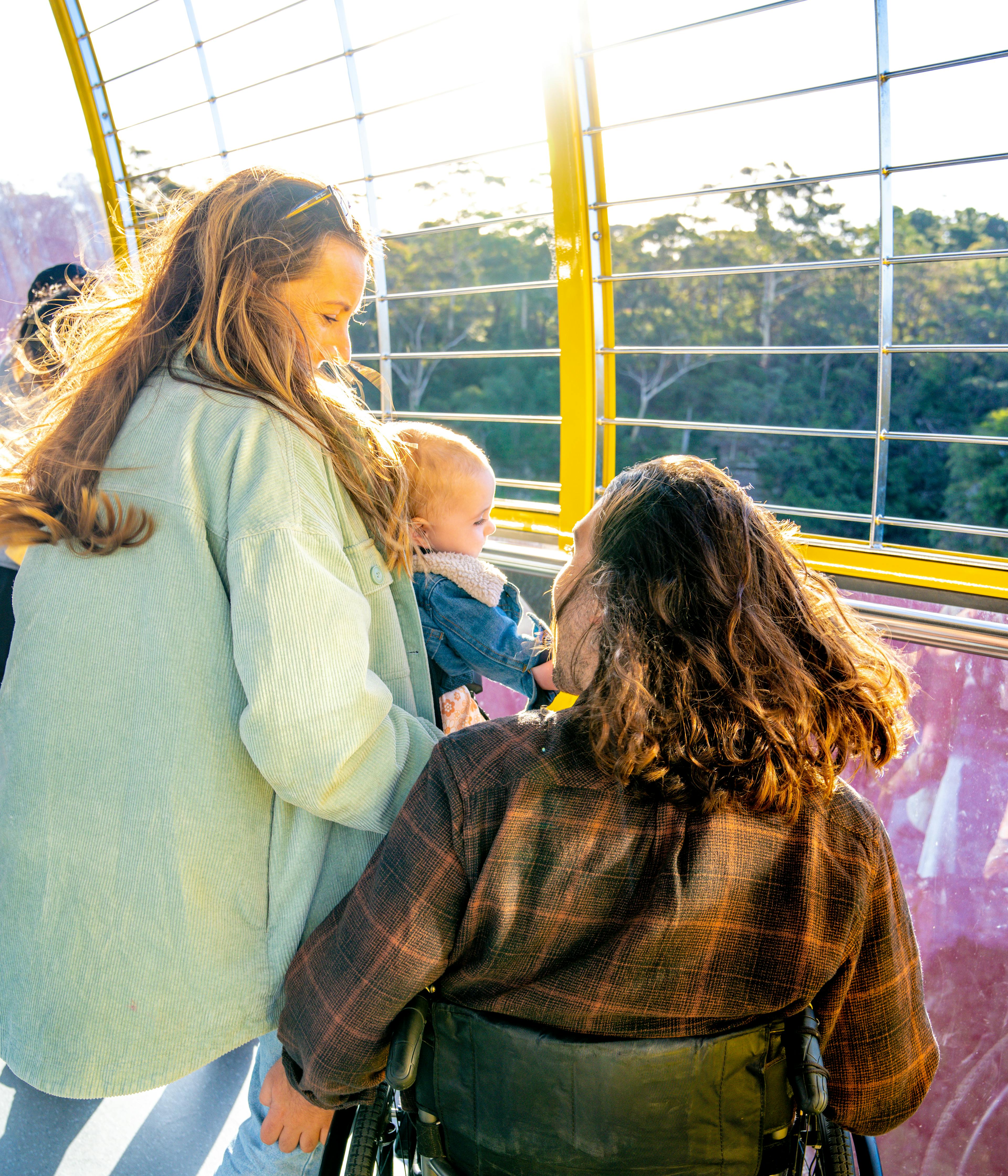 A family enjoys the view from within a cable car with large windows. A woman stands beside a man in a wheelchair, both smiling. The woman holds a baby, and sunlight streams through the window, creating a warm, bright atmosphere. Trees are visible outside.