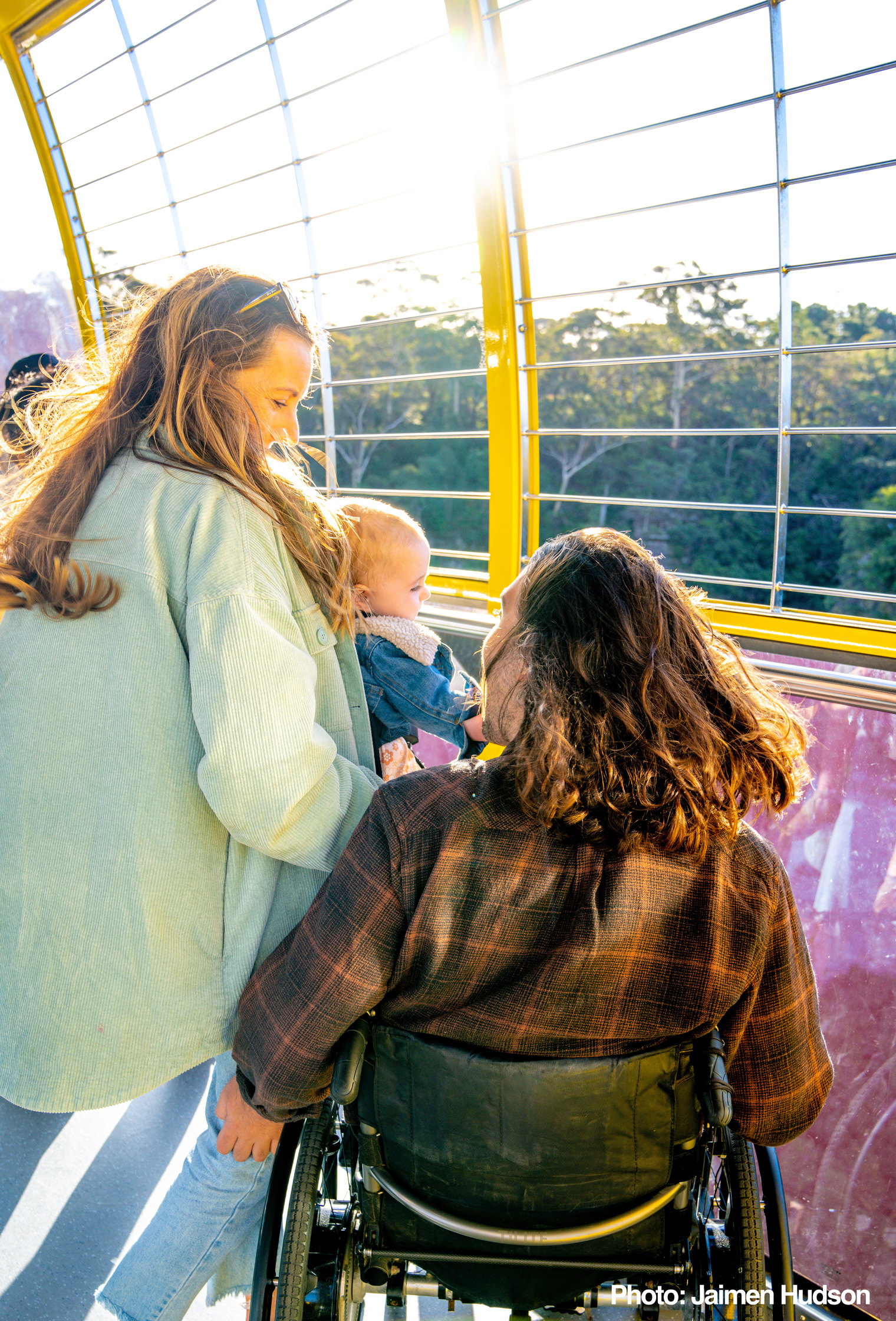 A couple with a baby enjoy a sunny day on an outdoor observation deck. The person in the wheelchair smiles at the baby while the other stands nearby. The background features trees and sunlight streaming through the railing.