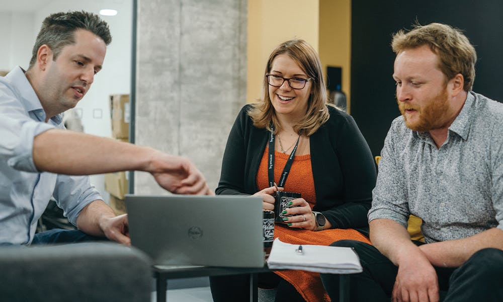 Three people in a meeting, with a person on the left pointing to a laptop screen