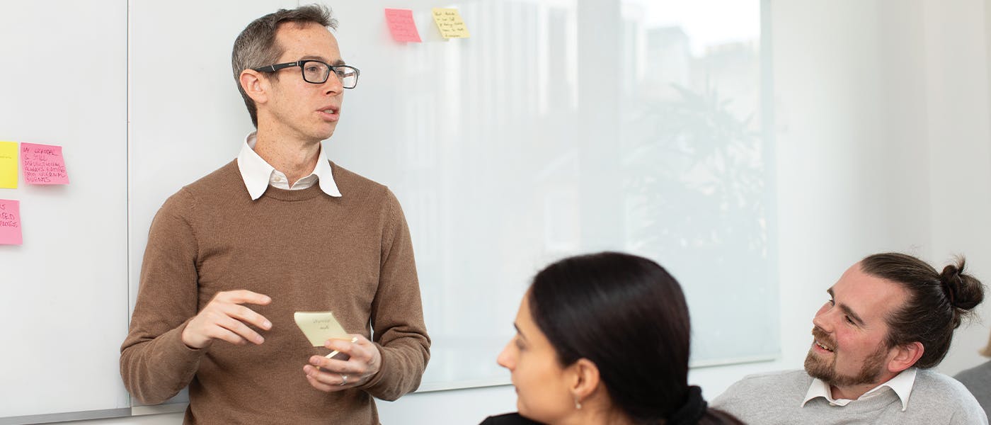 Person stands in front of white board with sticky notes addressing others