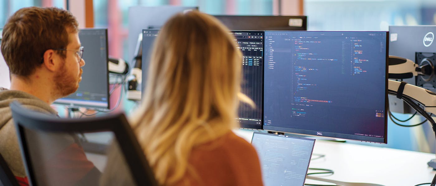 Two people sat at a desk, looking a computer monitor with code on it