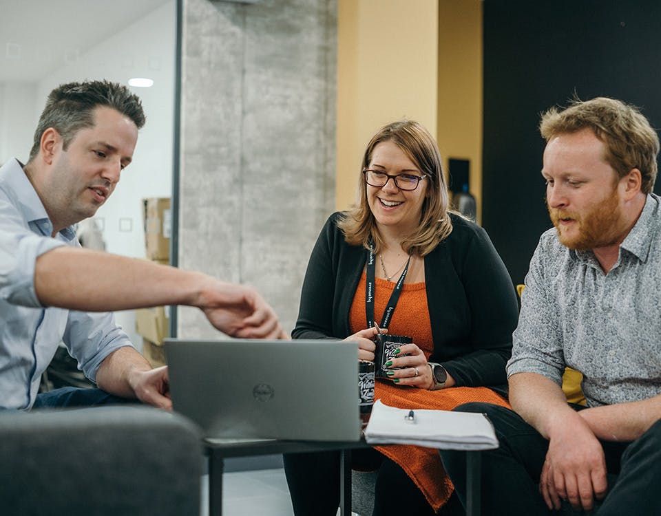 Three people in a meeting, with a person on the left pointing to a laptop screen
