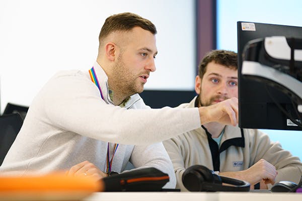 Two people at a desk looking at a computer screen, with one of them pointing at something on the screen