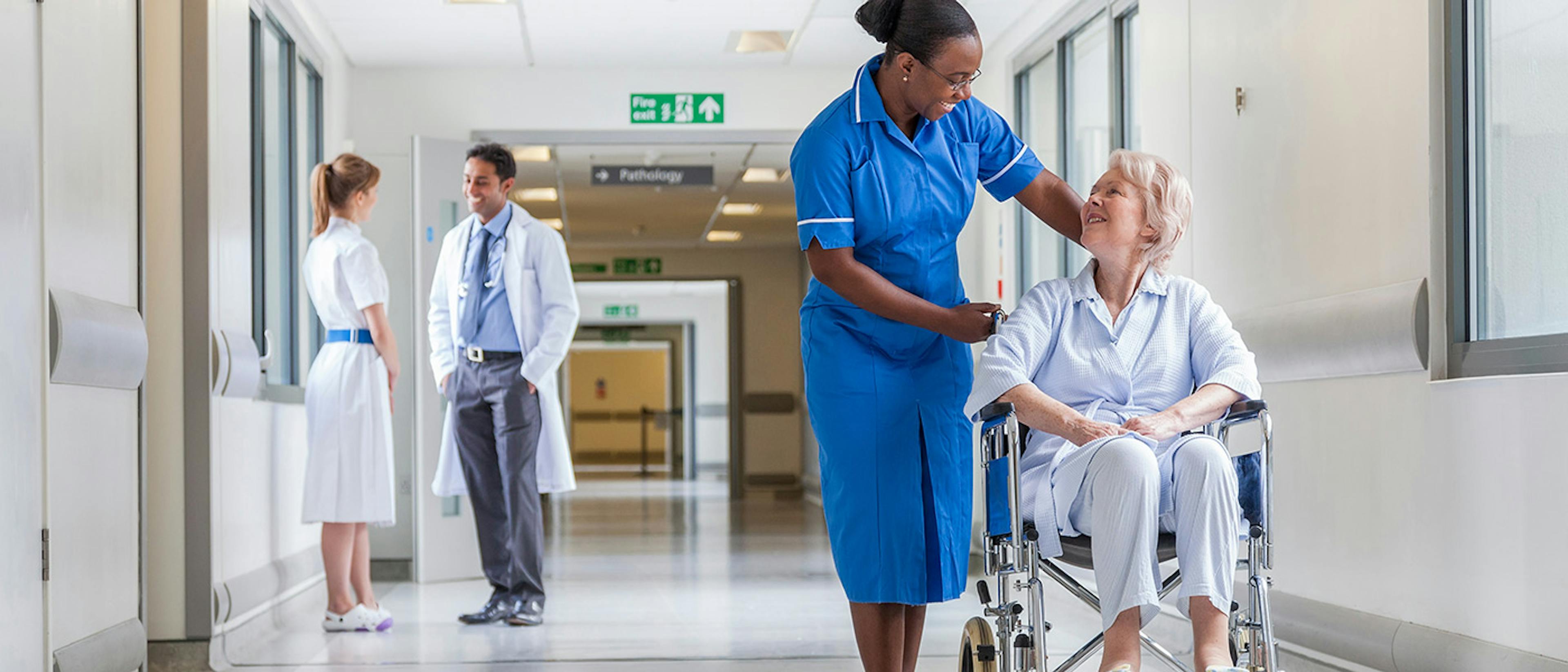 People in a hospital corridor, with a nurse and doctor talking on the left, and a nurse with a patient in a wheelchair on the right