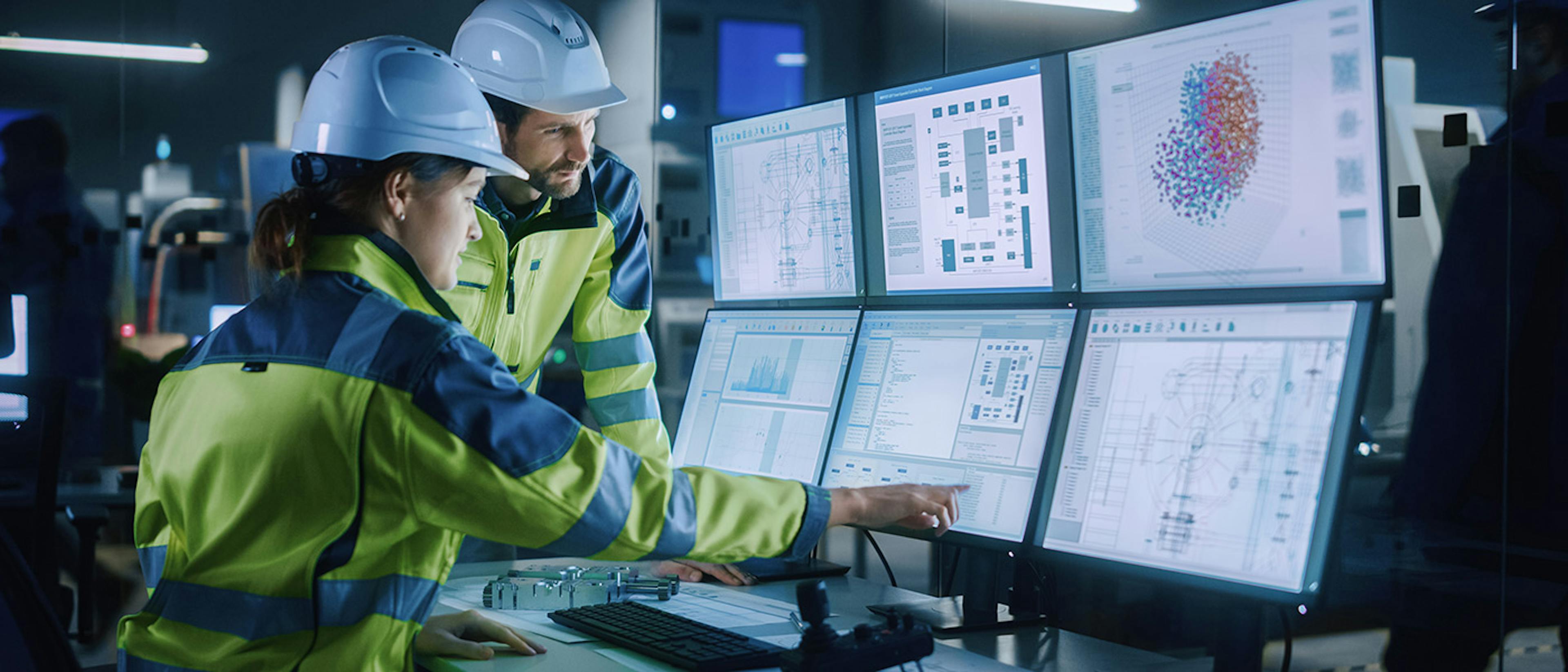 A woman and man in a factory with hi-vis coats and hard hats on, with a bank of screens in front of them, at which the woman is pointing.