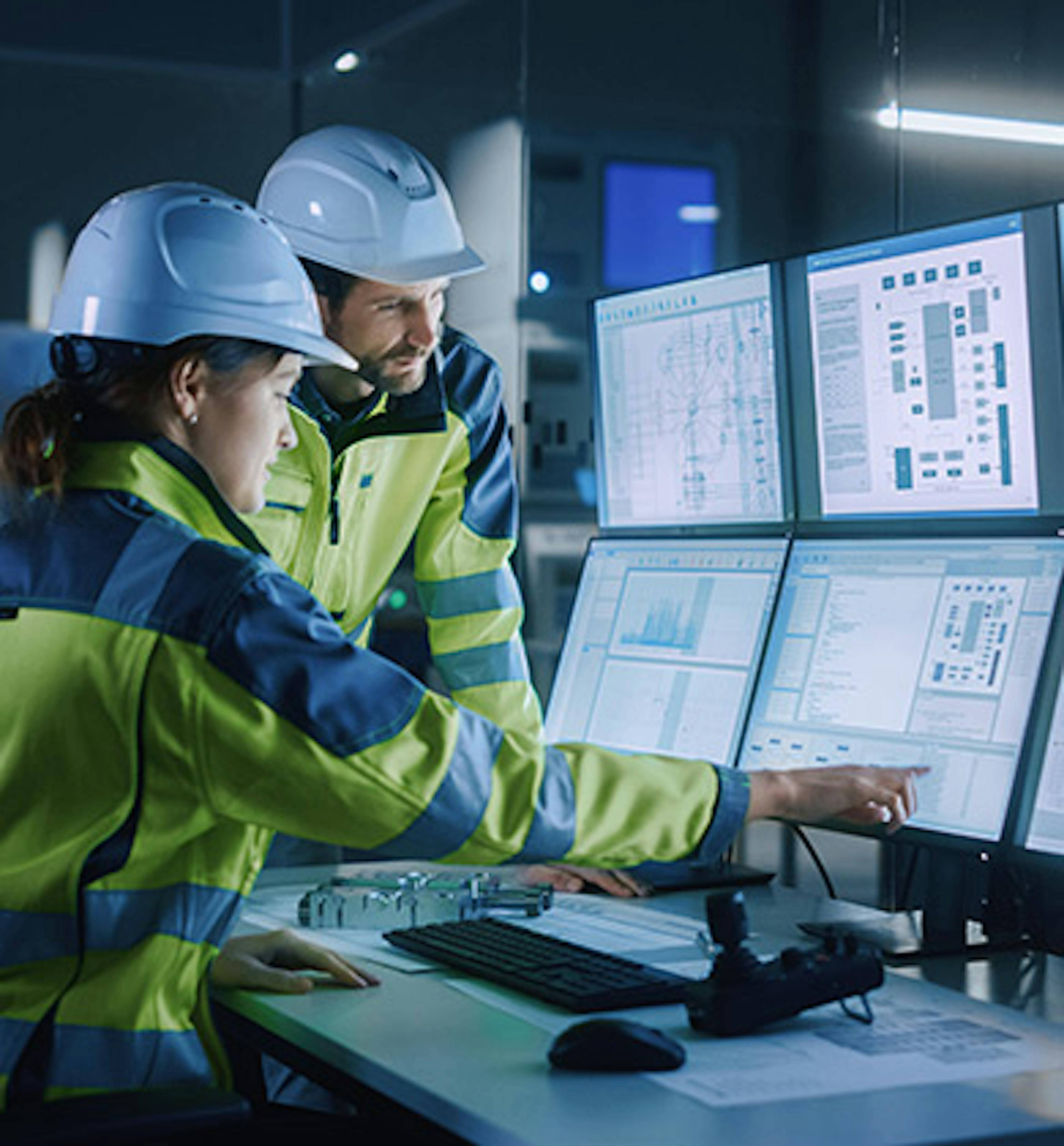 A woman and man in a factory with hi-vis coats and hard hats on, with a bank of screens in front of them, at which the woman is pointing.