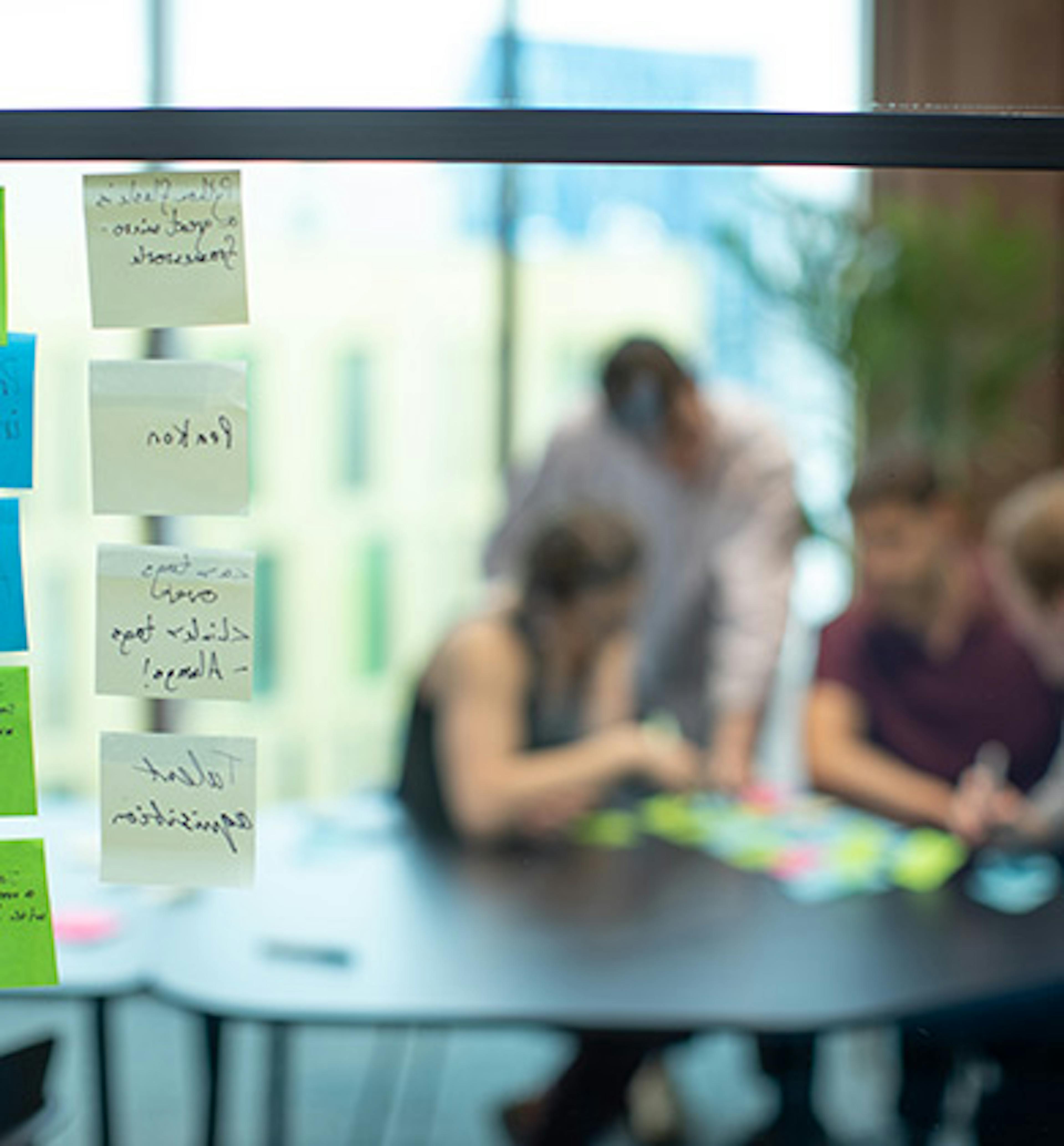 Sticky notes on a window on the left, and a blurred group of people on the right discussing other sticky notes