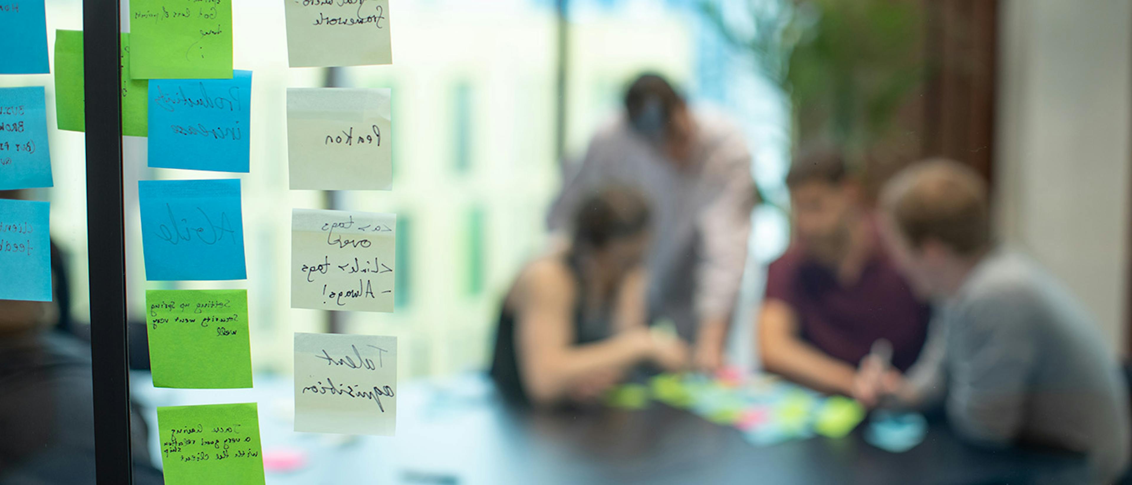 Sticky notes on a window on the left, and a blurred group of people on the right discussing other sticky notes