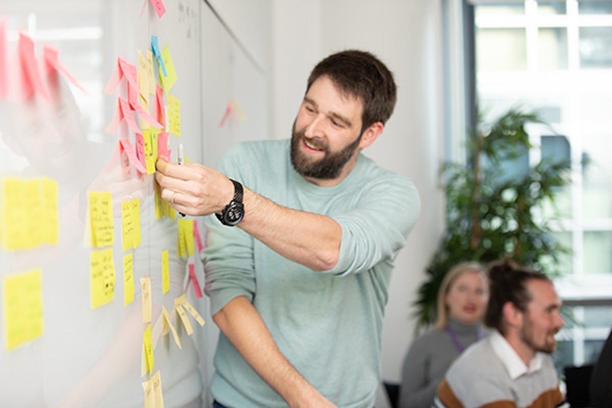 Person adding a sticky note to a whiteboard in a meeting, with colleagues in the background