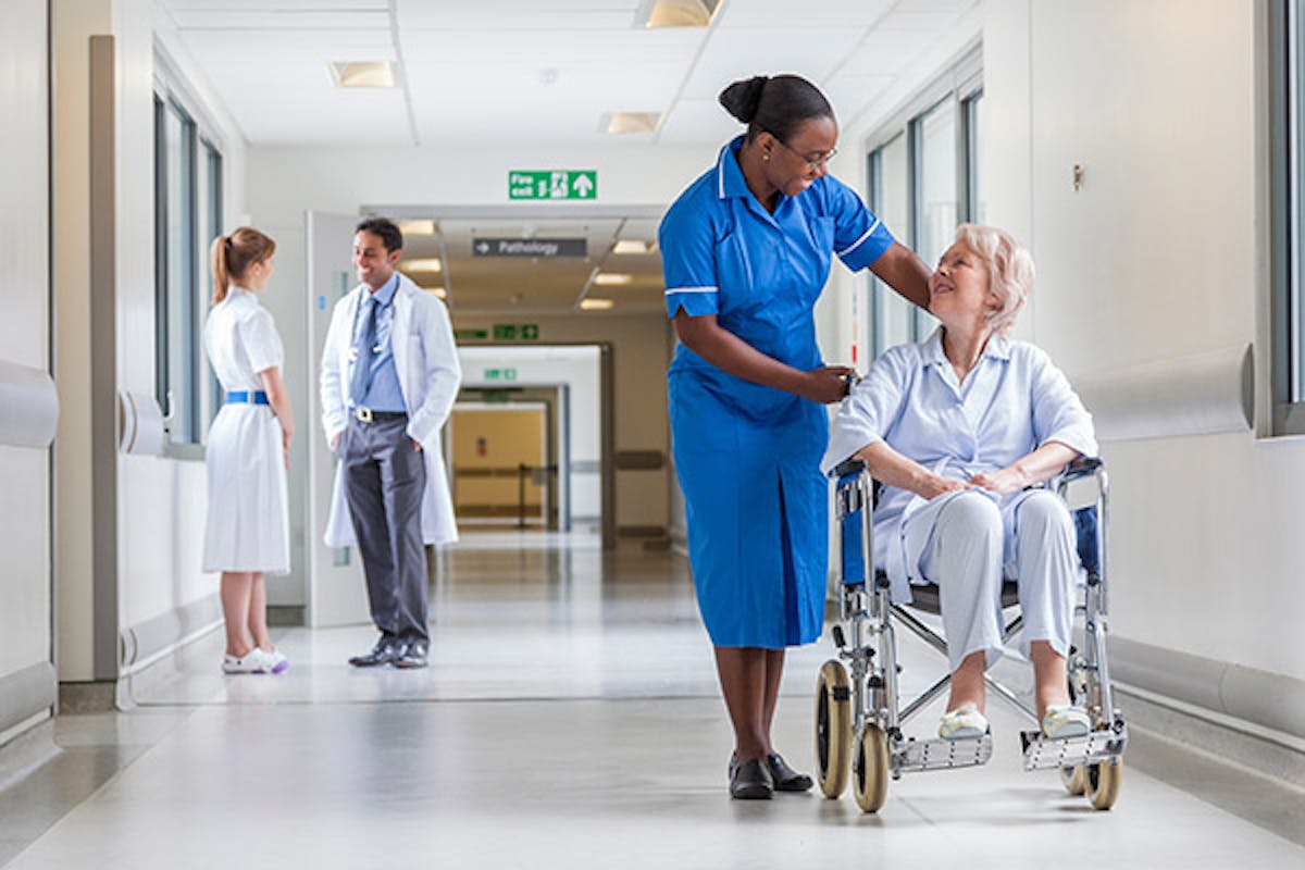 People in a hospital corridor, with a nurse and doctor talking on the left, and a nurse with a patient in a wheelchair on the right