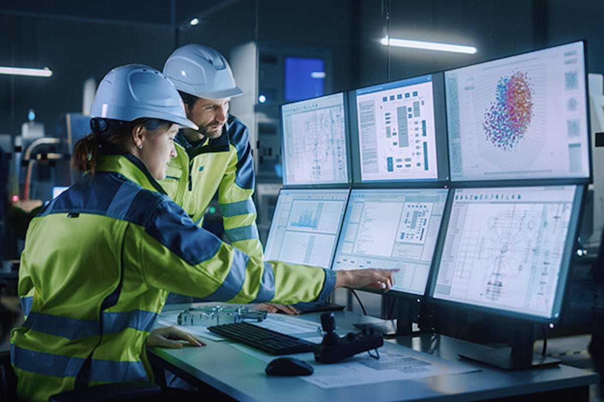 A woman and man in a factory with hi-vis coats and hard hats on, with a bank of screens in front of them, at which the woman is pointing.