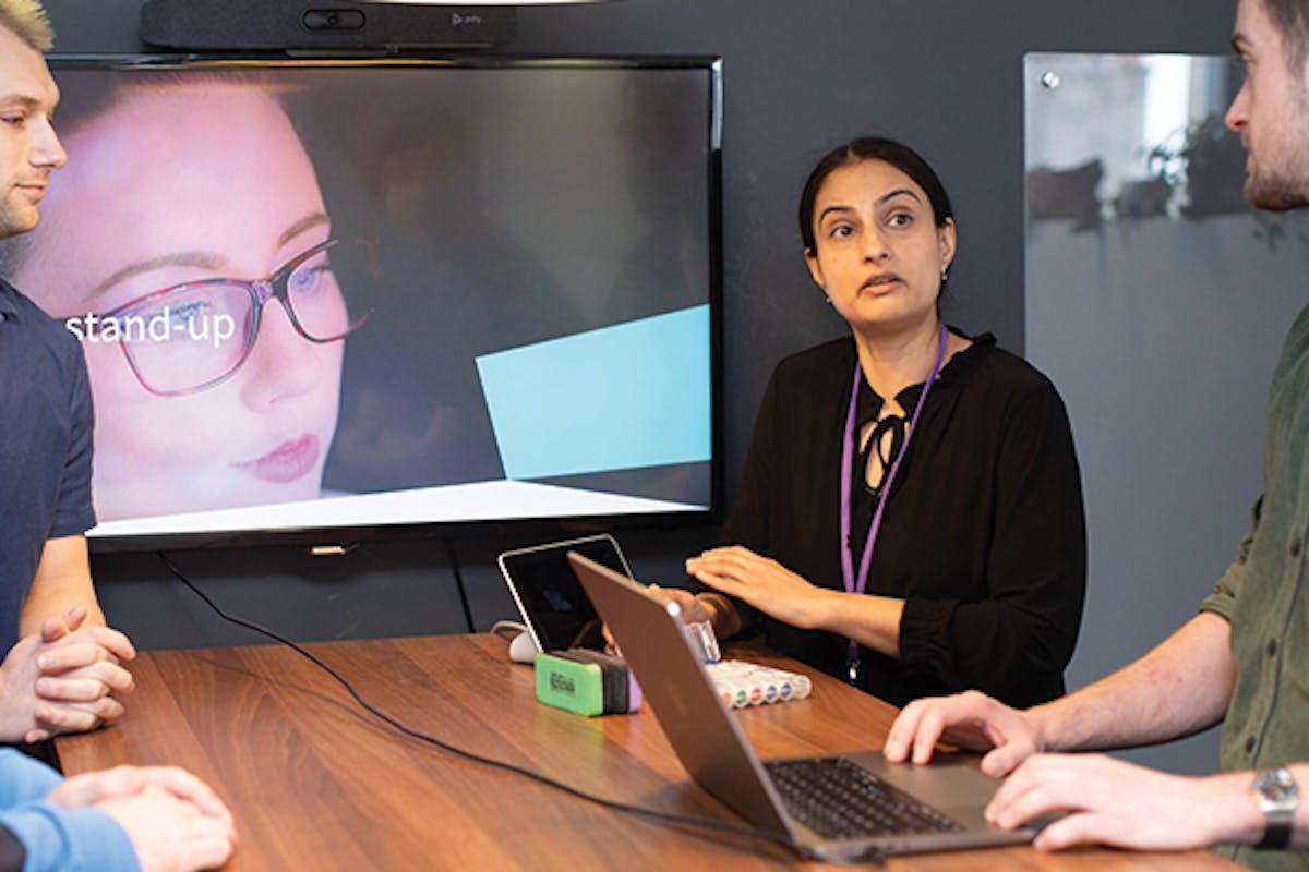 Employees gather around monitor and laptop for stand-up