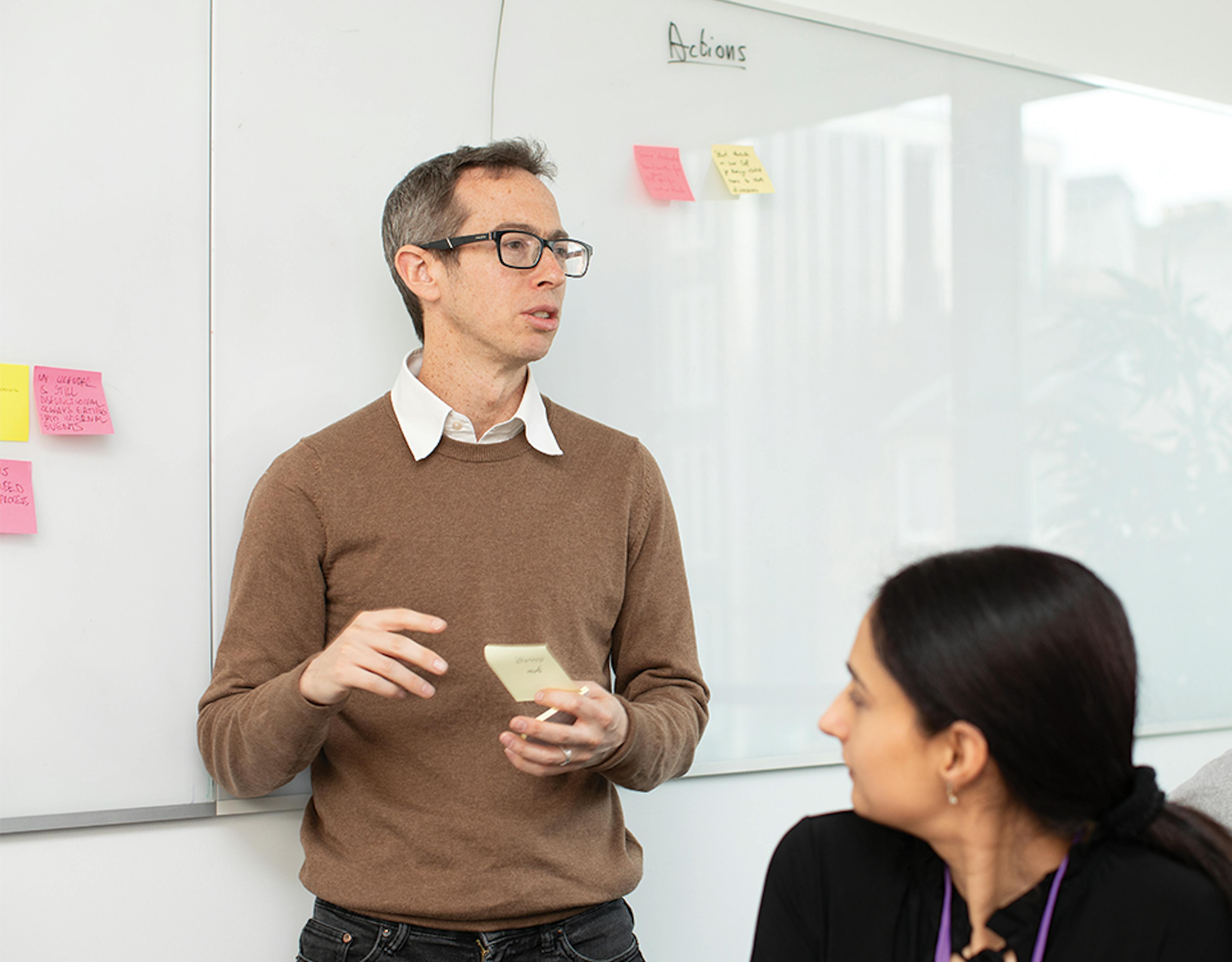 Person stands in front of white board with sticky notes addressing others