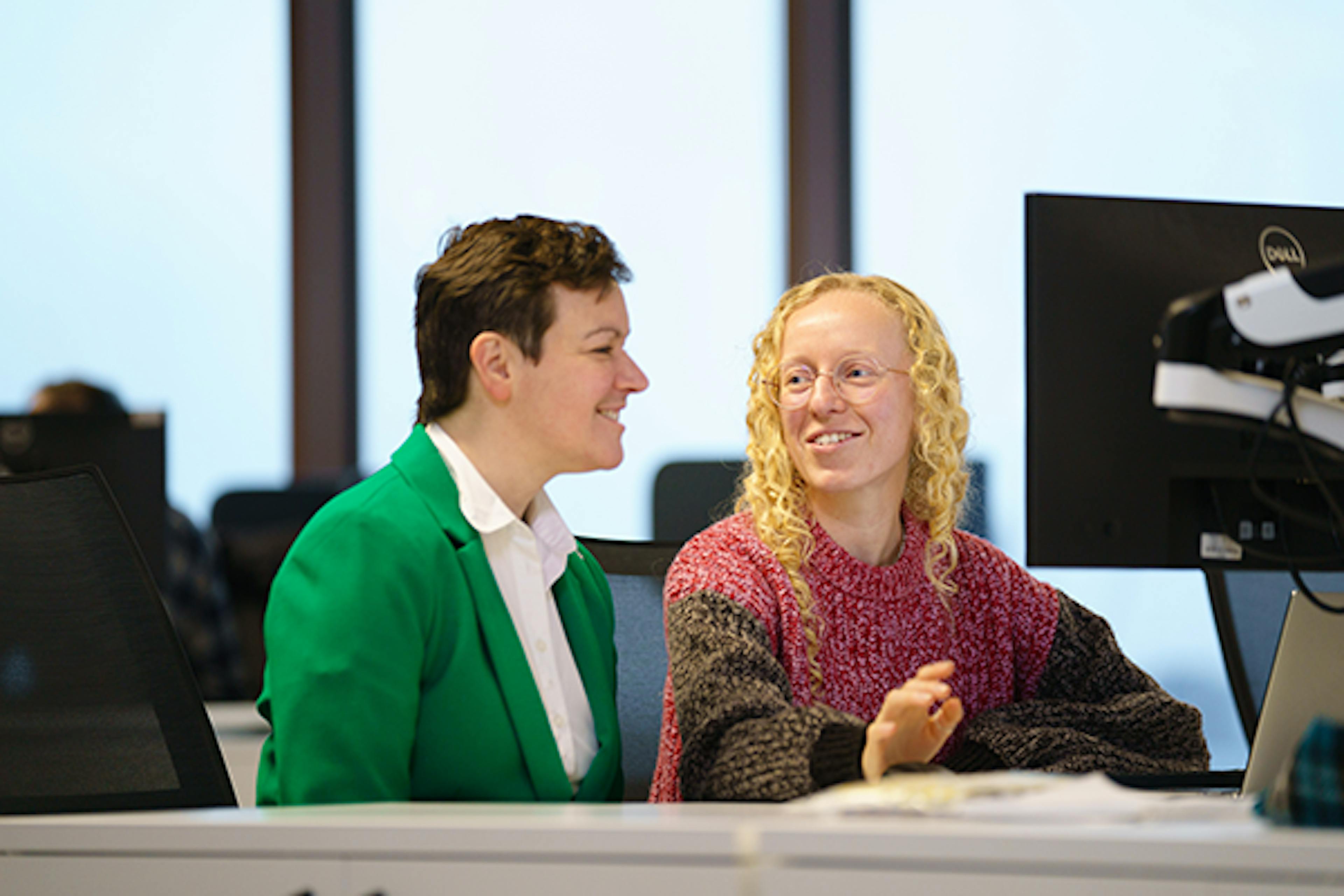 Two people talking at a desk in an open-plan office.