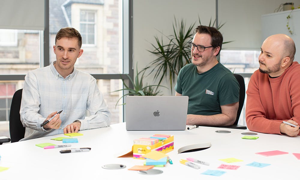 Three UX designers in a meeting around a big table with a laptop and sticky notes
