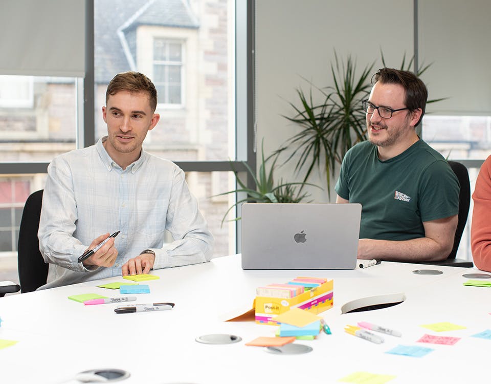 Three UX designers in a meeting around a big table with a laptop and sticky notes