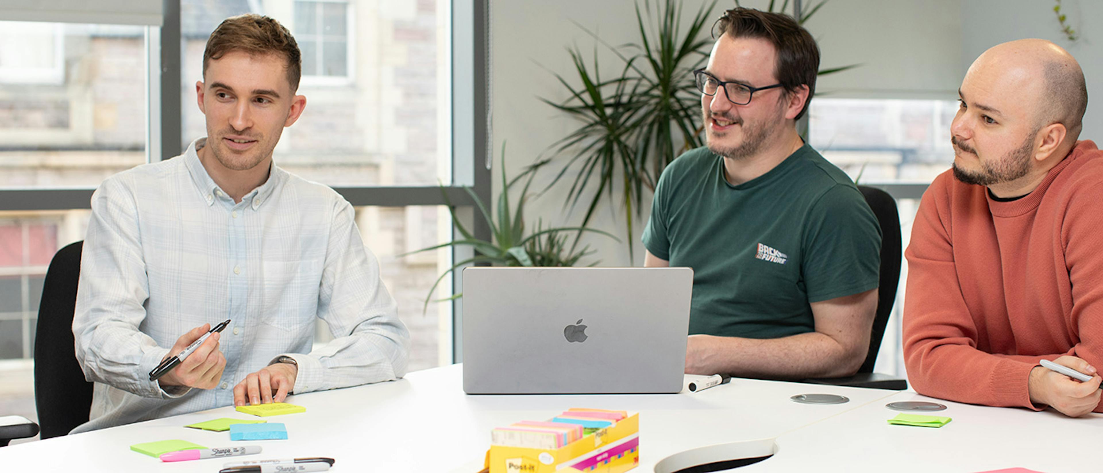 Three UX designers in a meeting around a big table with a laptop and sticky notes