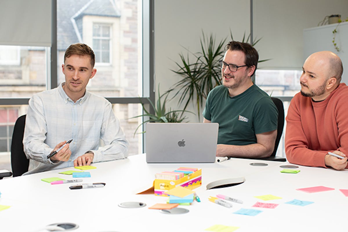 Three UX designers in a meeting around a big table with a laptop and sticky notes