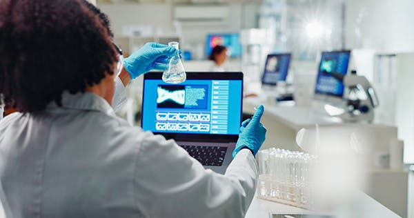 A person with their back to us, working in a lab and holding up a laboratory flask in front of a screen