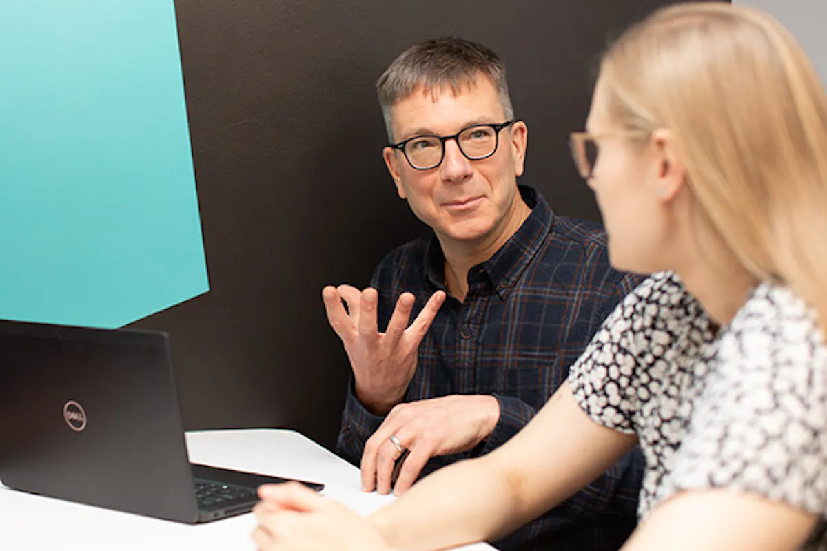 A person in an office meeting setting, sitting in front of a laptop and explaining something to their colleague.