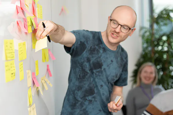 A person standing at a whiteboard in an office setting, talking while adding a sticky note to the board.