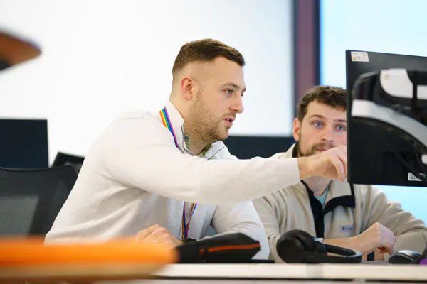 Two people at a desk looking at a computer screen, with one of them pointing at something on the screen