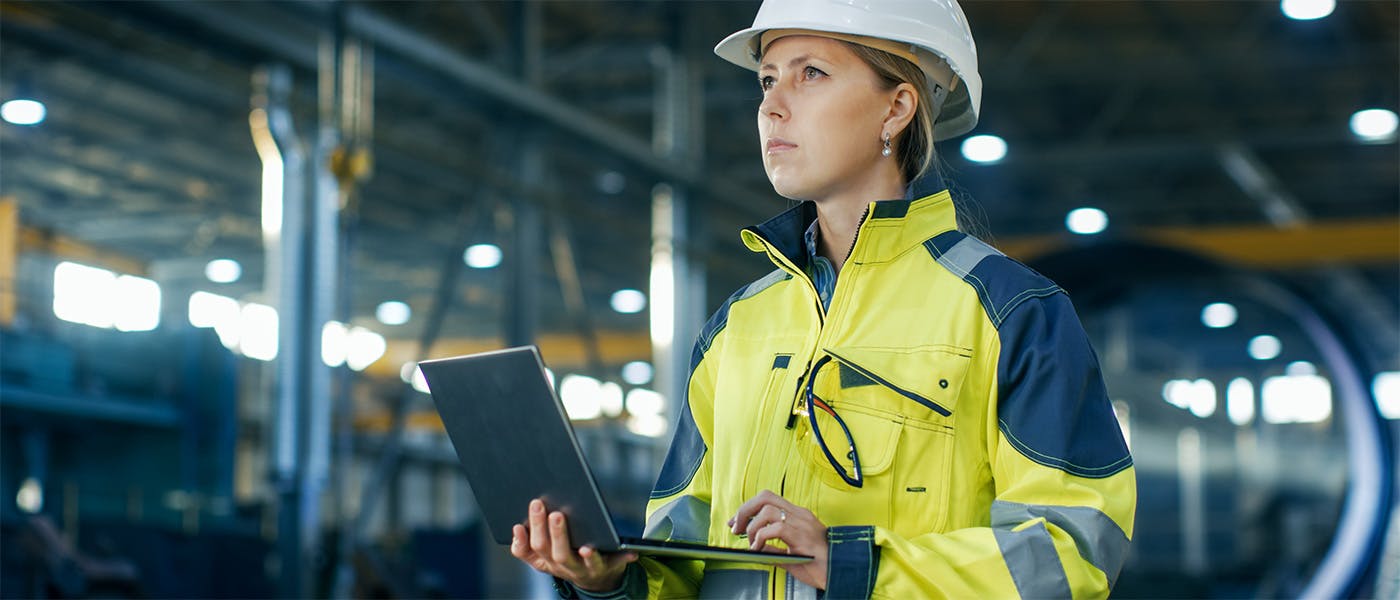 Woman in high-visibility coat and hard hat using a laptop on a production line