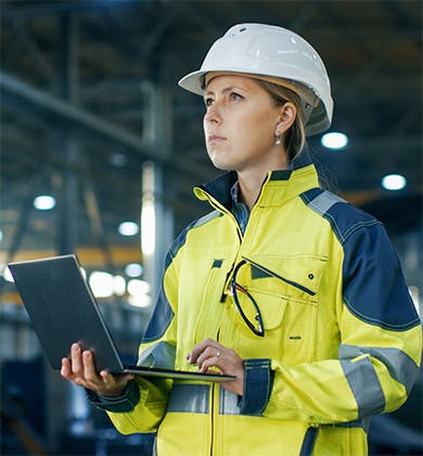 Woman in high-visibility coat and hard hat using a laptop on a production line