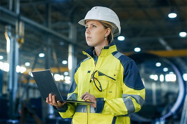 Woman in high-visibility coat and hard hat using a laptop on a production line