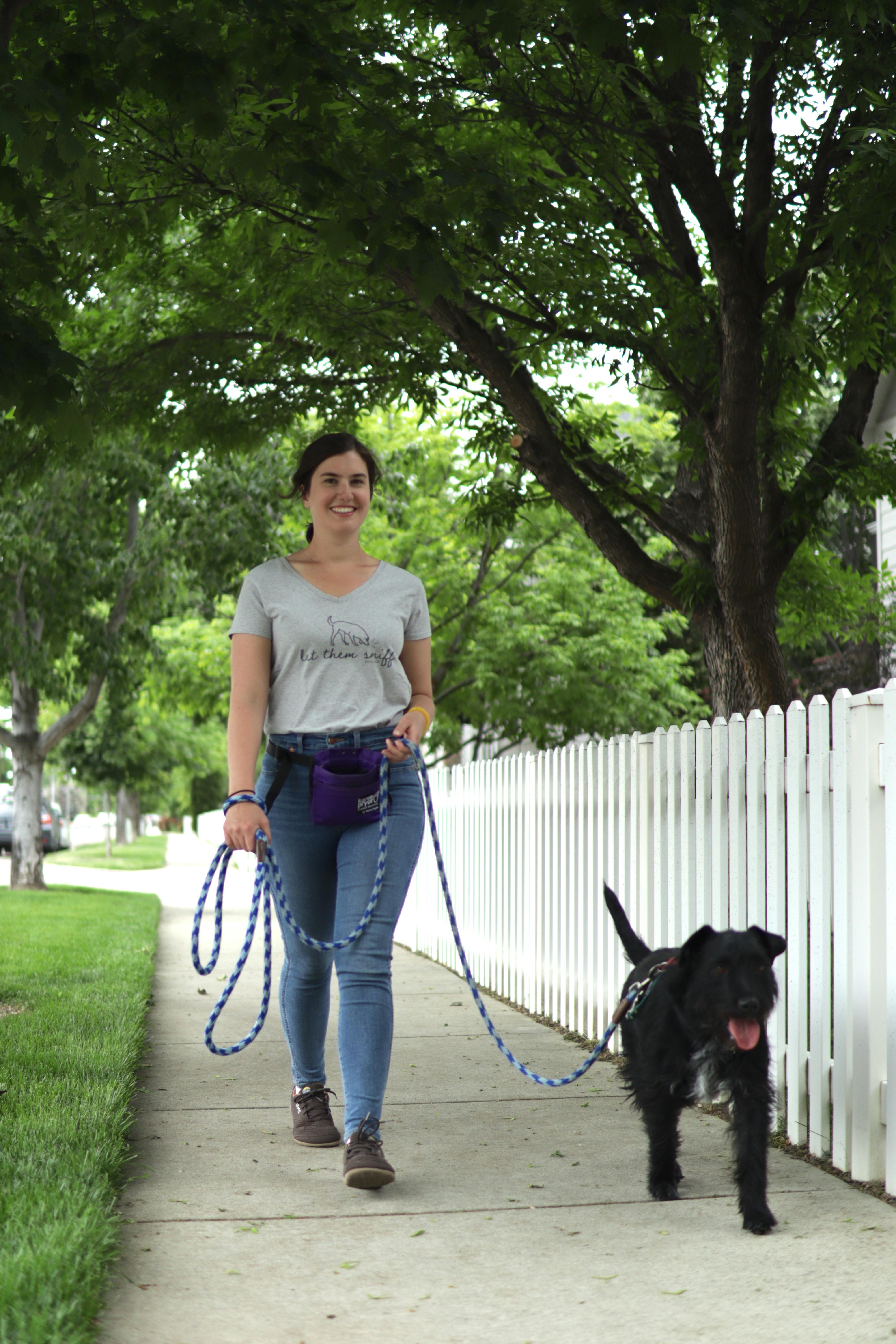 A woman walks a black dog on a long leash while smiling at the camera