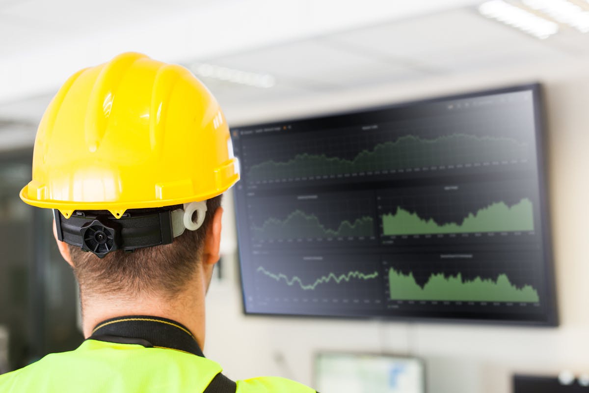 Man wearing a yellow hardhat and high-vis jacket looks at a screen showing data