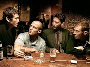 Four white men sat at a pub bar with drinks and cigarettes around them having an intent discussion