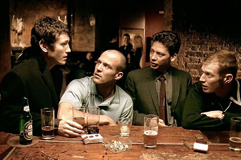 Four white men sat at a pub bar with drinks and cigarettes around them having an intent discussion