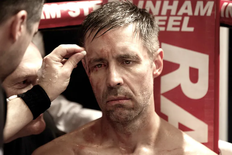 A scuffed up and sweaty man sitting in the corner of a boxing ring having his cuts attended to looking forlorn
