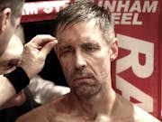 A scuffed up and sweaty man sitting in the corner of a boxing ring having his cuts attended to looking forlorn
