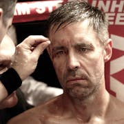 A scuffed up and sweaty man sitting in the corner of a boxing ring having his cuts attended to looking forlorn