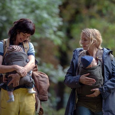 Two white women looking worn down, their babies strapped to their chests, carrying rucksacks as they walk through a woods