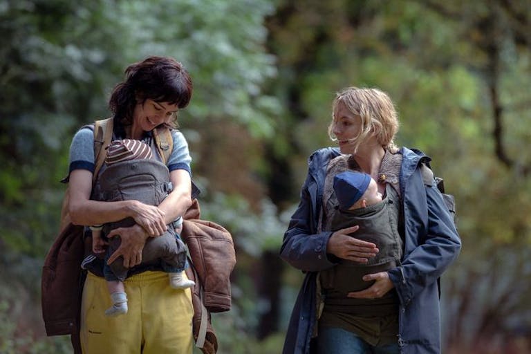 Two white women looking worn down, their babies strapped to their chests, carrying rucksacks as they walk through a woods